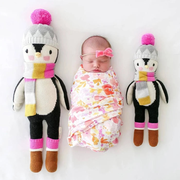 Newborn baby lying between two Cuddle + Kind Aspen the Penguin dolls, one large and one small, wearing colorful scarves and winter hats.