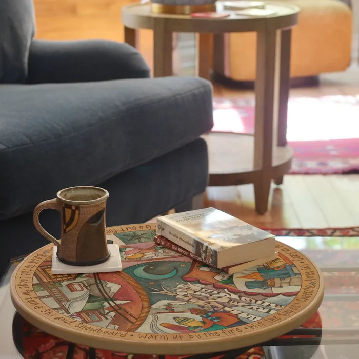 A round wooden coffee table with a colorful illustrated design and carved lazy susan sits in a living room setting. A ceramic mug and a book rest on the table, which is placed in front of a blue armchair.