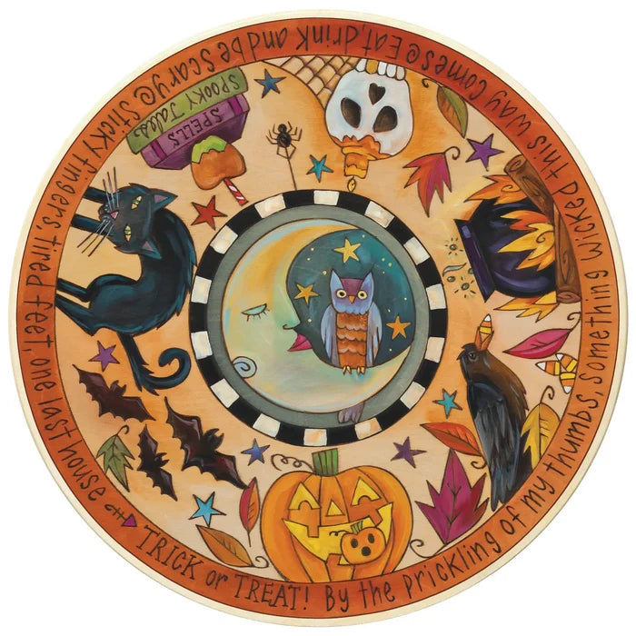 A circular Halloween-themed decorative lazy susan with a whimsical painted design. The center features an owl under stars inside a black-and-white checkered ring, surrounded by a warm orange border with illustrations of a black cat, a jack-o'-lantern, a raven, autumn leaves, stars, and handwritten text around the edge.