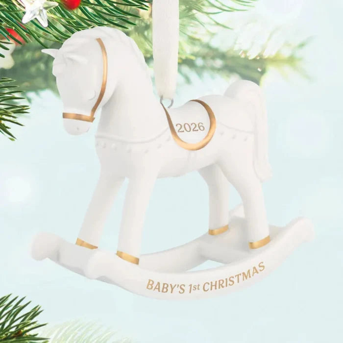 White rocking horse ornament with gold accents, '2026' and 'BABY'S 1ST CHRISTMAS' text, on a Christmas tree.