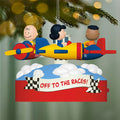 Decorative Christmas ornament with cartoon characters on a race car, 'Off to the Races!' text, and checkered flags against a blurred Christmas tree background.