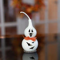 A white ceramic Halloween gourd decoration with a ghost face, an orange bow tie, and bat cutouts, displayed on a reflective dark surface with a blurred indoor background.