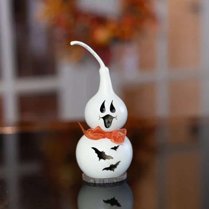 A white ceramic Halloween gourd decoration with a ghost face, an orange bow tie, and bat cutouts, displayed on a reflective dark surface with a blurred indoor background.