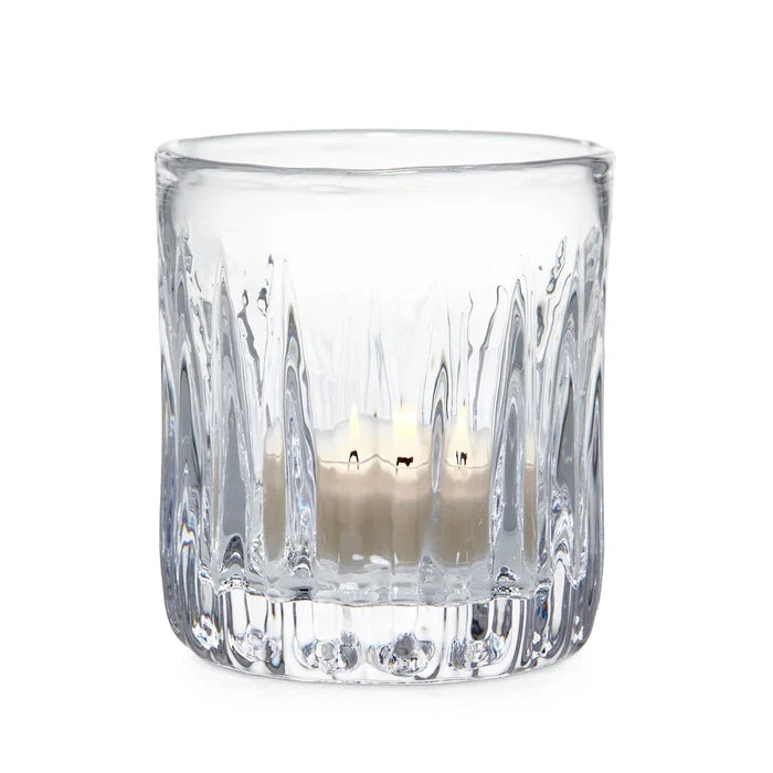 A clear glass candle holder containing two lit candles, casting a warm glow in a softly lit environment.