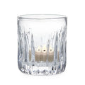 A clear glass candle holder containing two lit candles, casting a warm glow in a softly lit environment.