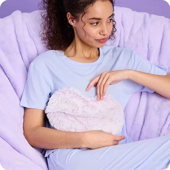 Woman holding a fluffy heart-shaped pillow in a cozy setting with purple bedding.